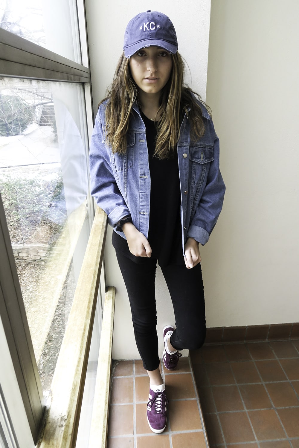 Kansas City native Hannah wears a black tee with black cuffed jeans, an oversized denim menswear jacket, and a blue KC hat. To add a pop of color, she wears magenta Adidas sneakers with her relaxed look.
