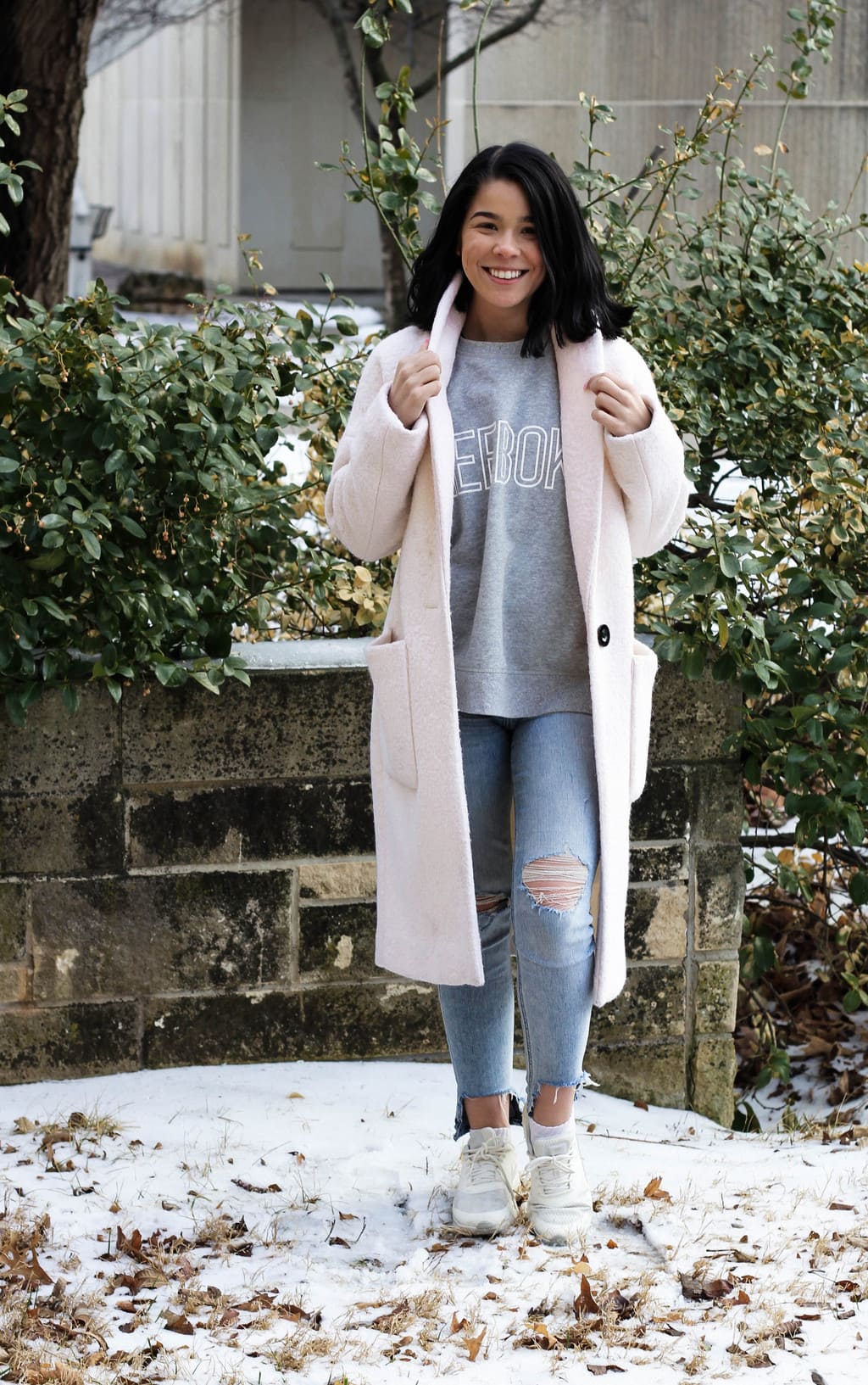 Kansas State University student Jessie sports a basic grey Reebok tee with distressed light-wash skinny jeans, white sneakers, and a long, baby pink coat.