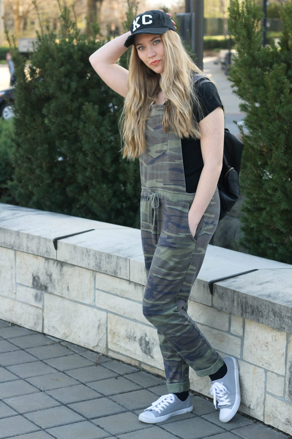 Monica, a student at Kansas State University, sports a relaxed knit camoflauge pair of overalls with a simple black tee, a black and white KC ballcap, a black backpack, and grey sneakers.