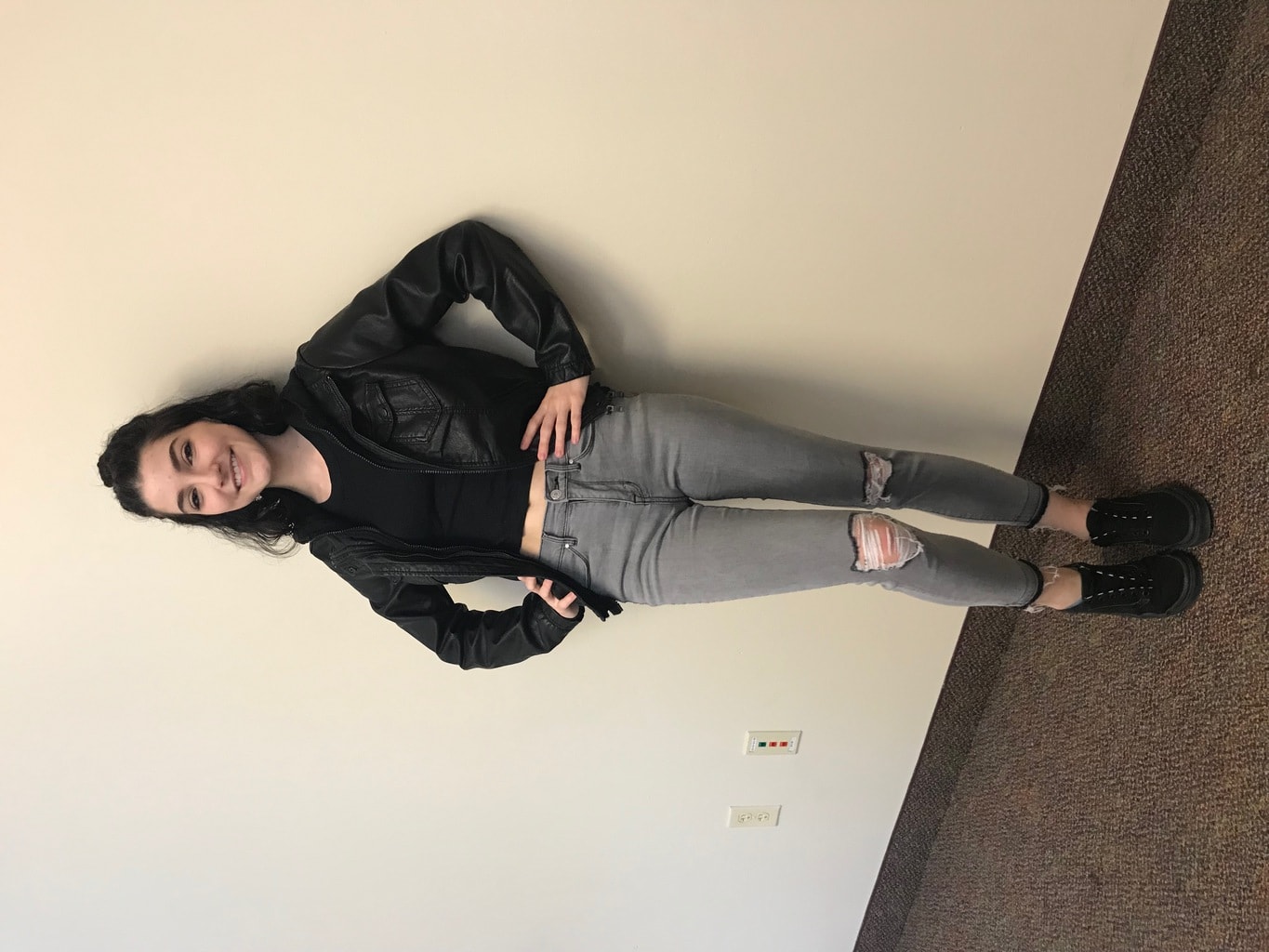Mercyhust University student Cara sports a black and grey ensemble made up of a black cropped tee and layered motorcycle jacket with a hood, grey distressed denim jeans, and flat black Vans sneakers.