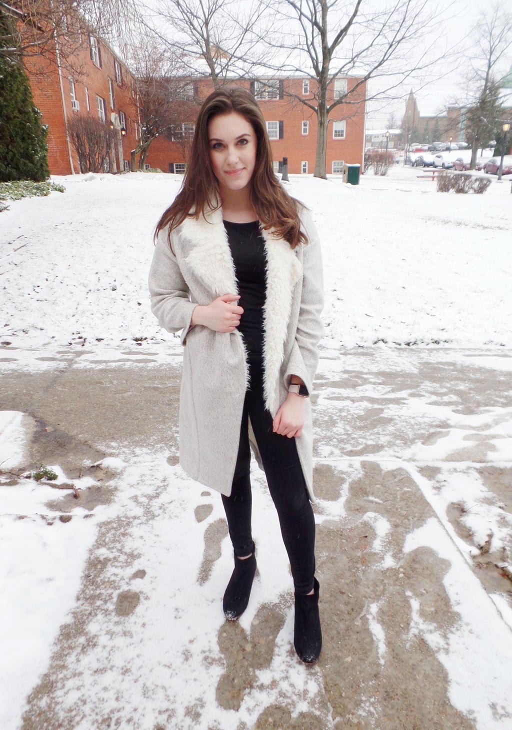 A Mercyhurst University student wears an all black monochromatic outfit made up of a tee, jeans, and booties, with a long grey coat with white fur lapels.