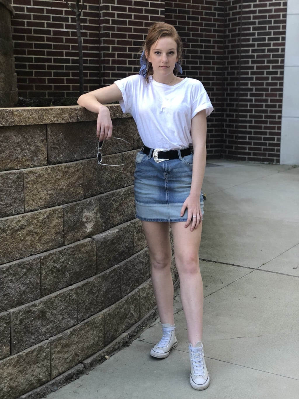 Emily wears a loose-fitted plain white tee tucked into a two-toned denim high-waisted skirt with a black belt and a thick silver western buckle. She pairs it with a blue bandana tied around her ponytail and high-top light grey converse sneakers.