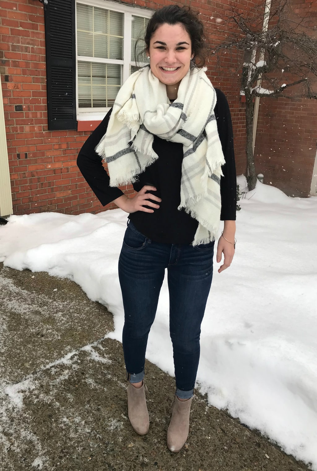 Erica wears a black 3/4-length sleeve tee with a chunky white and grey plaid scarf, dark wash cuffed skinny jeans, and taupe ankle booties.