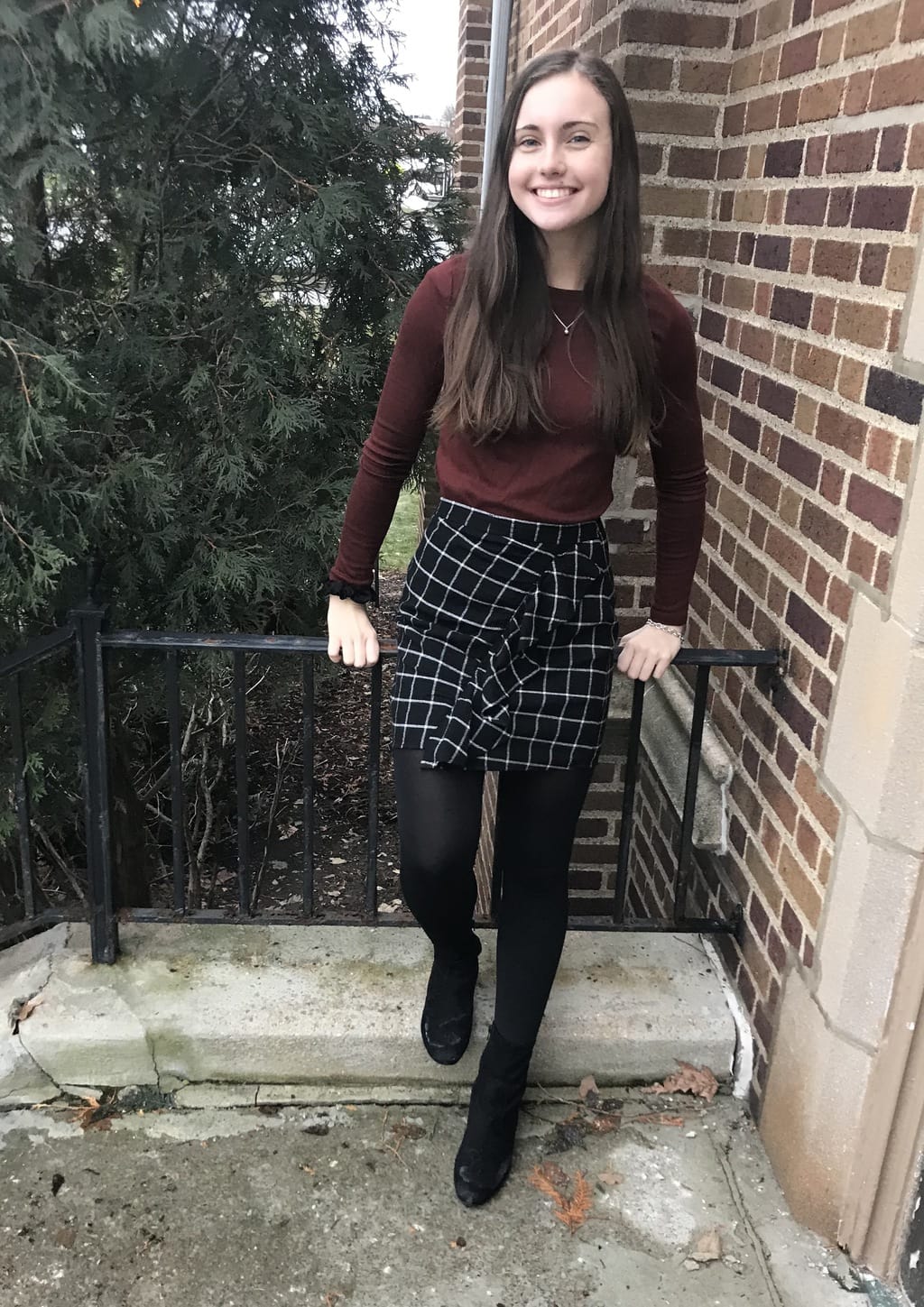 Mercyhurst University student Jillian looks put-together and professional in a maroon long-sleeve shirt with a black and white plaid ruffled skirt, black tights, and chunky black ankle booties.