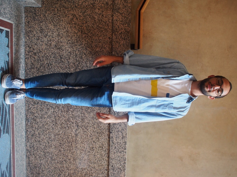 Andrea, a student at Politecnico di Milano, wears a white, yellow, blue, and blush graphic tee with a chambray denim shirt over. He pairs it with skinny blue jeans and scuffed up blue vans with white laces.