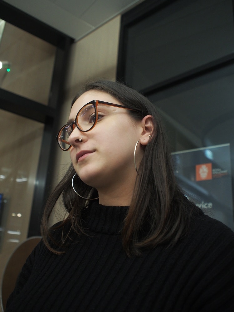 Dina wears a dainty silver nosering, silver hoop earrings, and black plastic glasses.