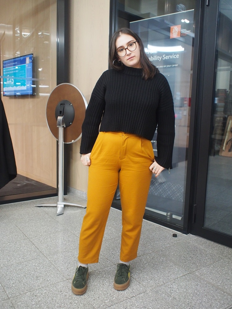 Royal Holloway student Dina wears a chunky black ribbed crop sweater with zippers on the sleeves, high-waisted mustard trousers, and chunky hunter green sneakers.