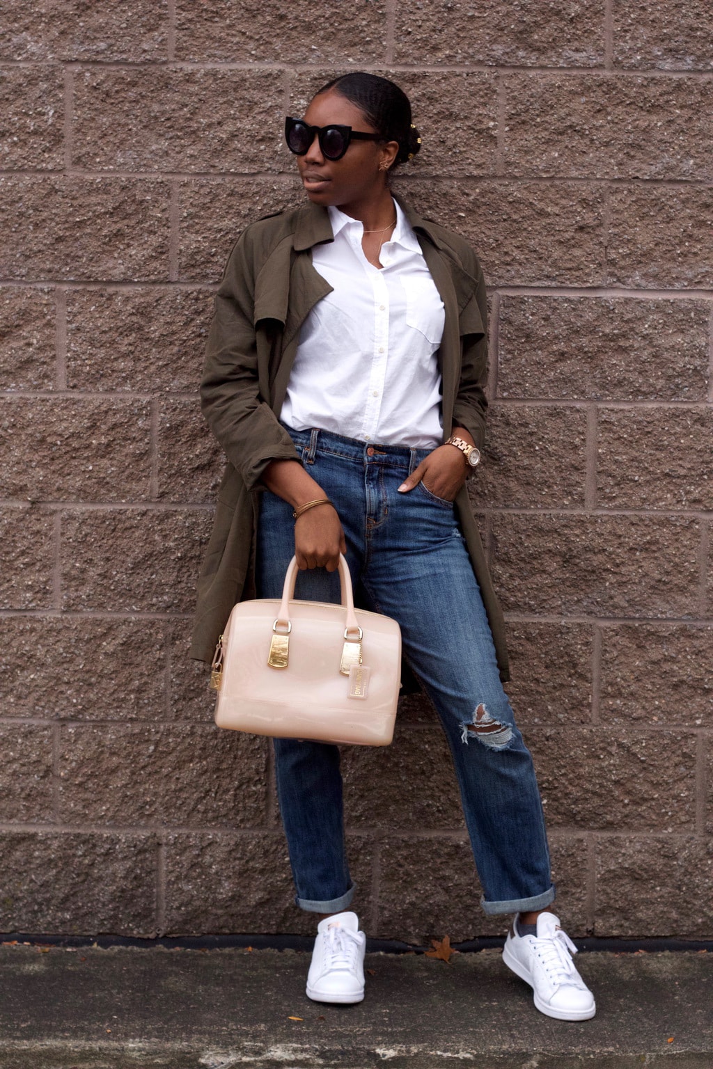 Amani wears a white button-up blouse tucked into loose-fit medium-wash distressed boyfriend jeans with an olive green duster rain jacket and white sneakers. She accessorizes with a blush pink rubberized tote bag and bold cat-eye sunglasses.