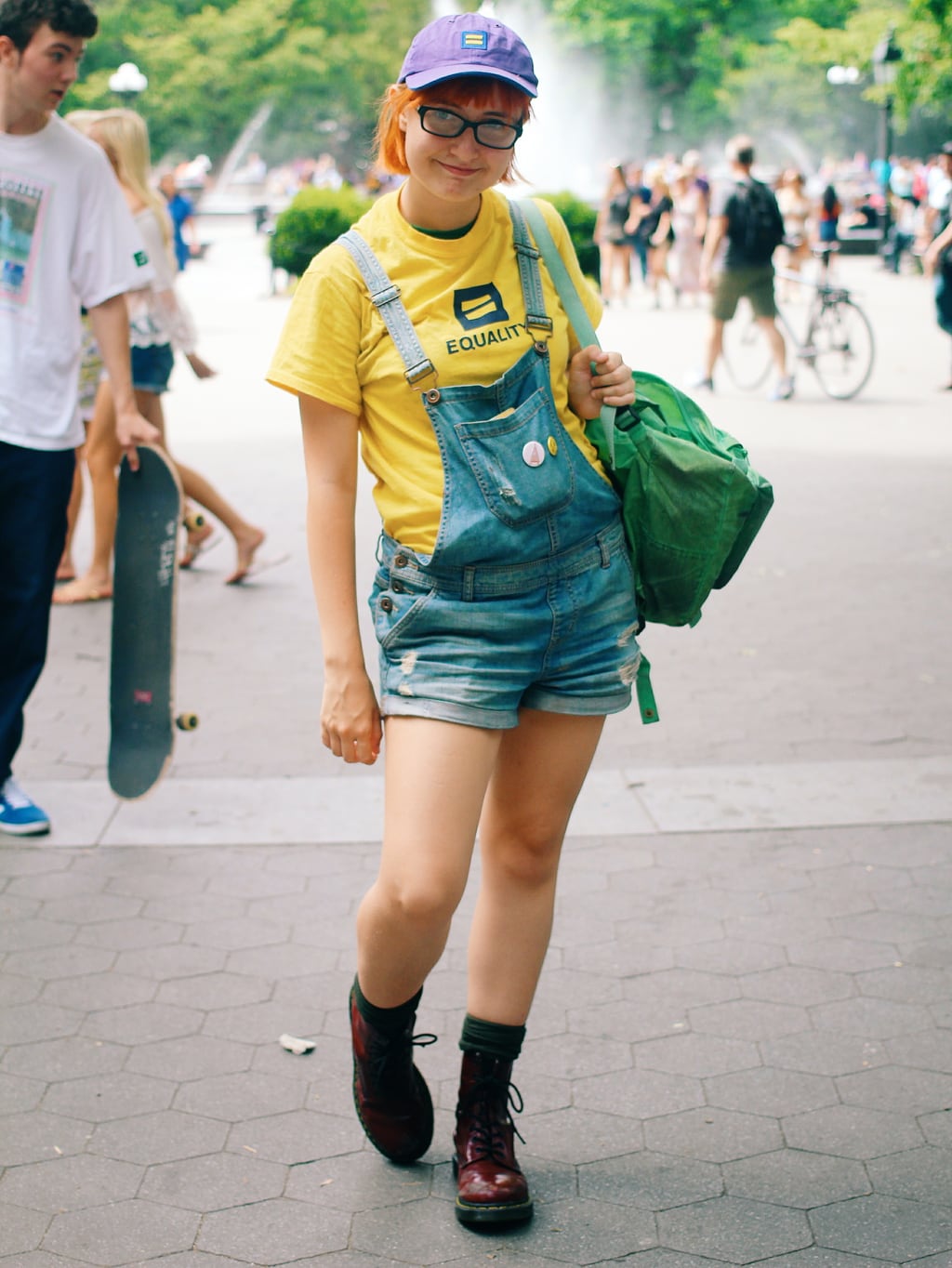 Fashion at School of Visual Arts, NYC - student Kaylee wears a quirky summer outfit idea with an Equality hat and tee shirt, distressed overalls, a green backpack, and maroon Doc Martens boots