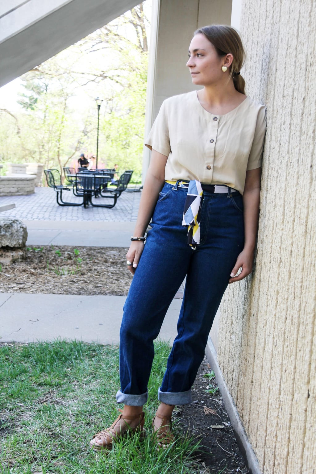 Emilee, a student at Kansas State University, wears a pale yellow scoop neck button-up short sleeve top with brown buttons, high-waisted deep blue denim jeans, a yellow, blue, and black vintage scarf tied as a belt, brown woven sneakers, and heart-shaped earrings.