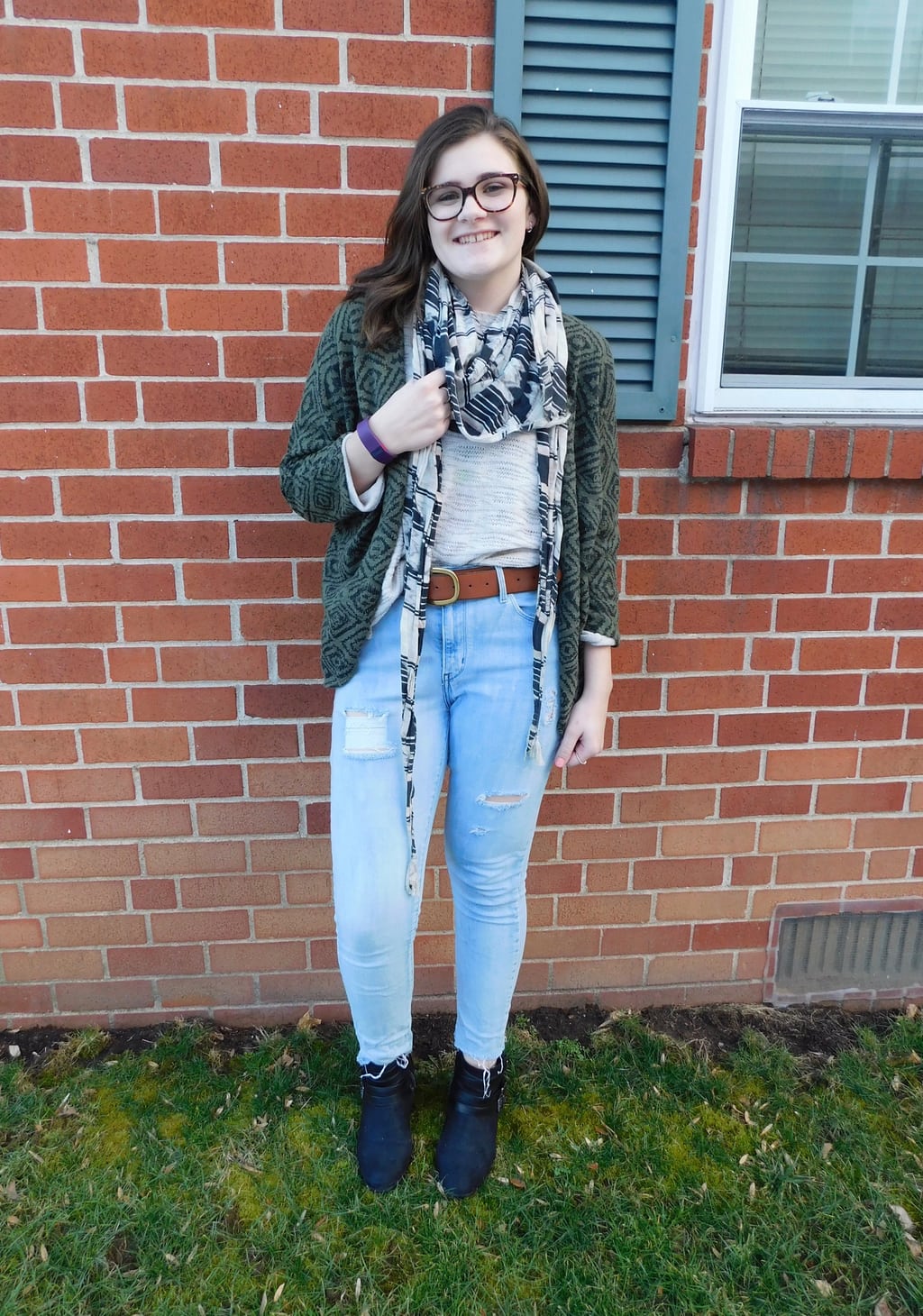 Lauren sports a cream sweater top with an olive green geometric print cardigan and a brown belt on her distressed denim skinny jeans. She tops off her look with a bold black and white scarf and black booties.