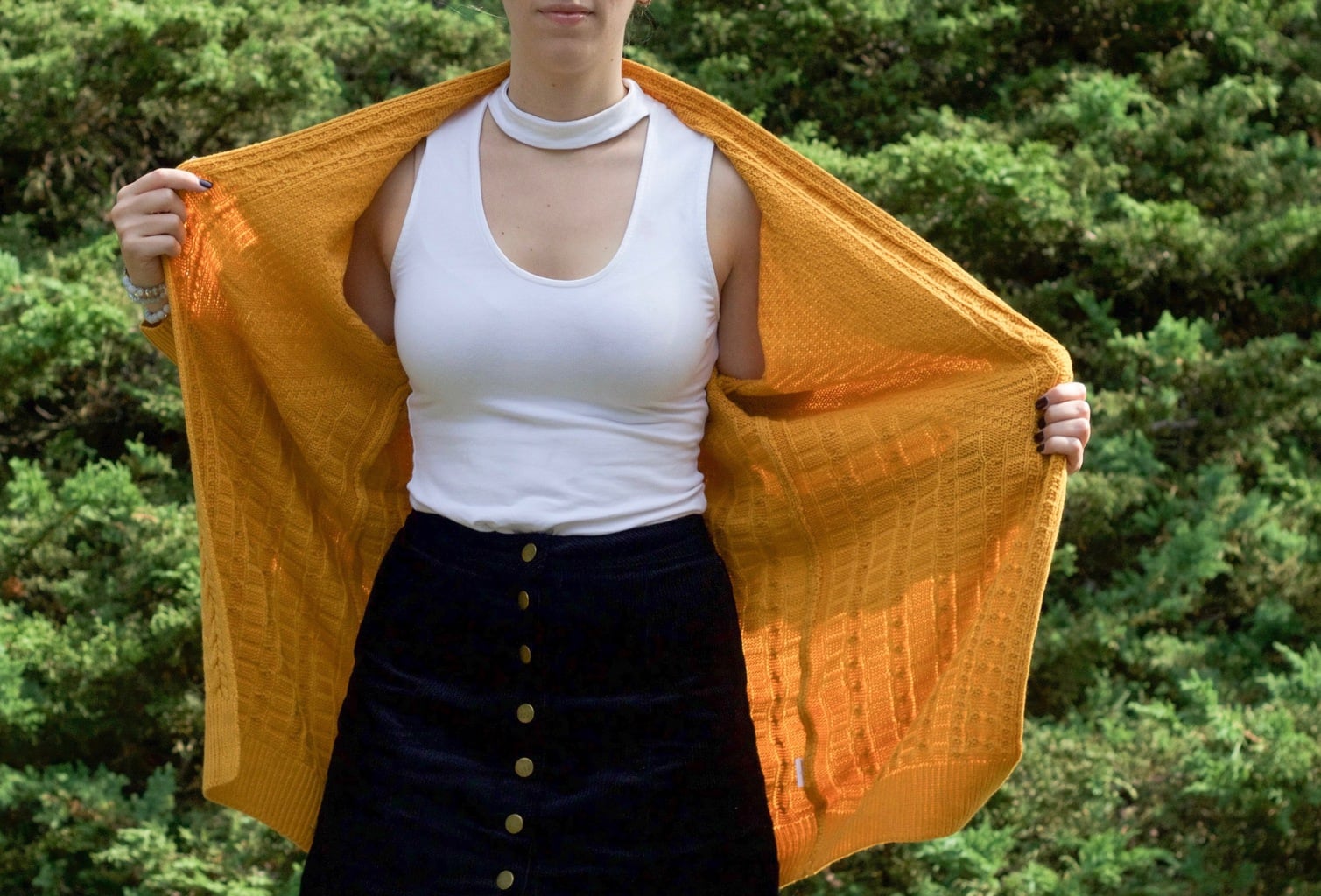 Edgy white tank top with mockneck and chest cutout, paired with a yellow knit cardigan and button-up skirt.