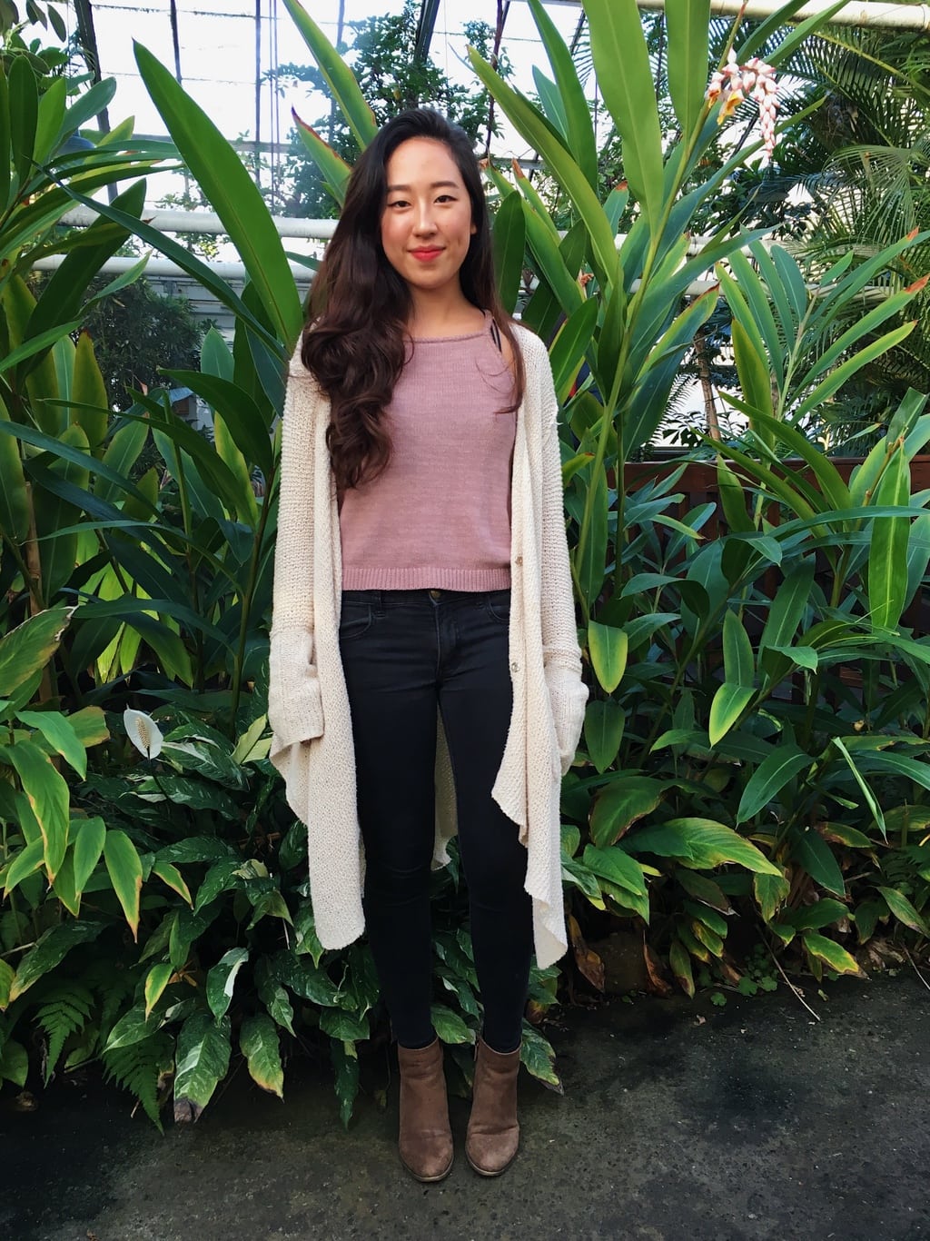 University of Massachusetts Amherst student Aren sports a mauve knit sleeveless top, a long flowy oatmeal knit sweater, dark wash skinny jeans, and chunky brown booties in the campus greenhouse.