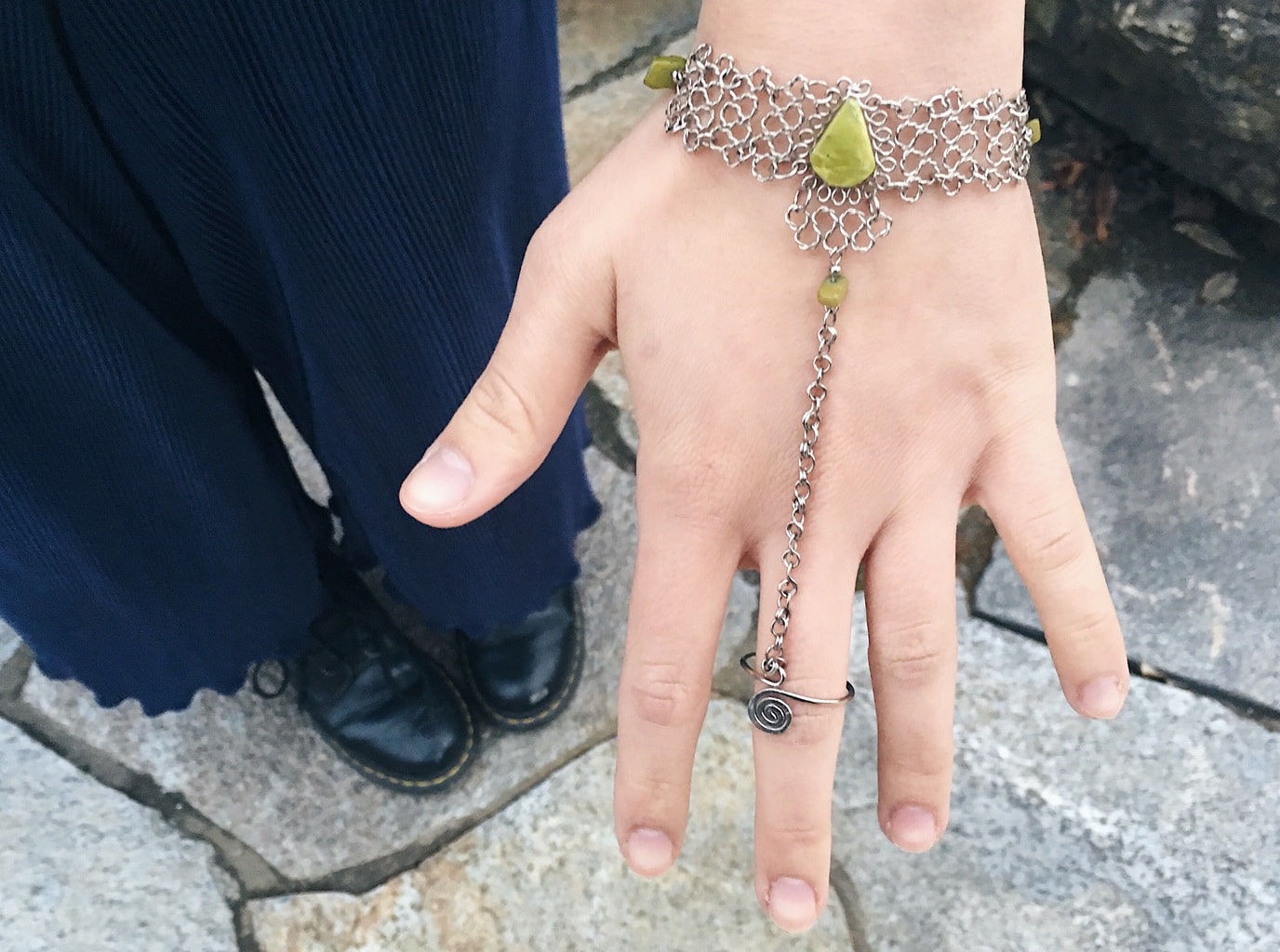 Caeli wears unique silver hand bracelet with detailed metal links that connect a ring to a bracelet. This piece has yellow-green stones to accent.