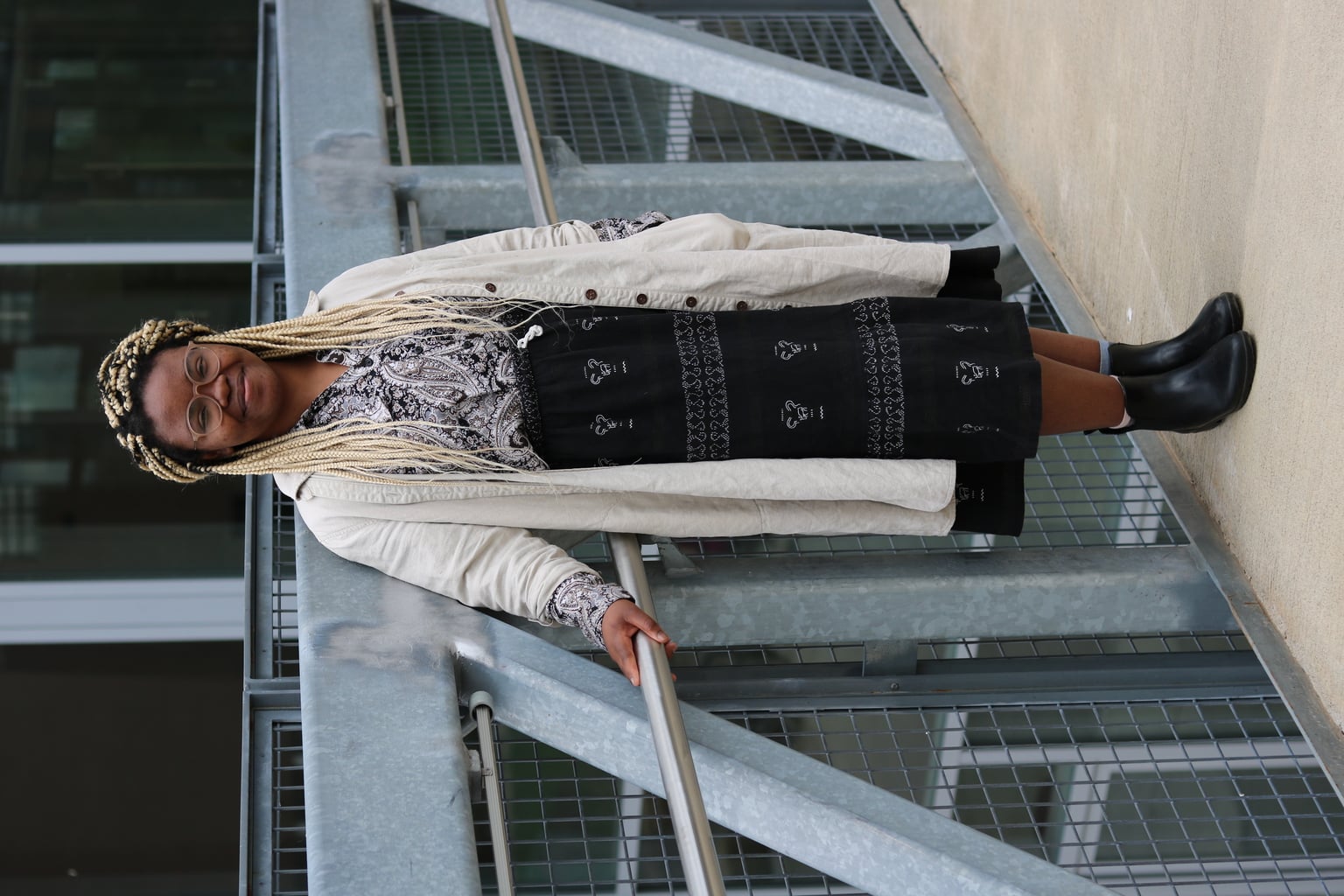 UMass Amherst student Cynthia wears a white and black long sleeve paisley button-up collared shirt tucked into a flowy bohemian midiskirt with delicate white stitching. She pairs it with a long canvas tan trench coat and flat rain boots.