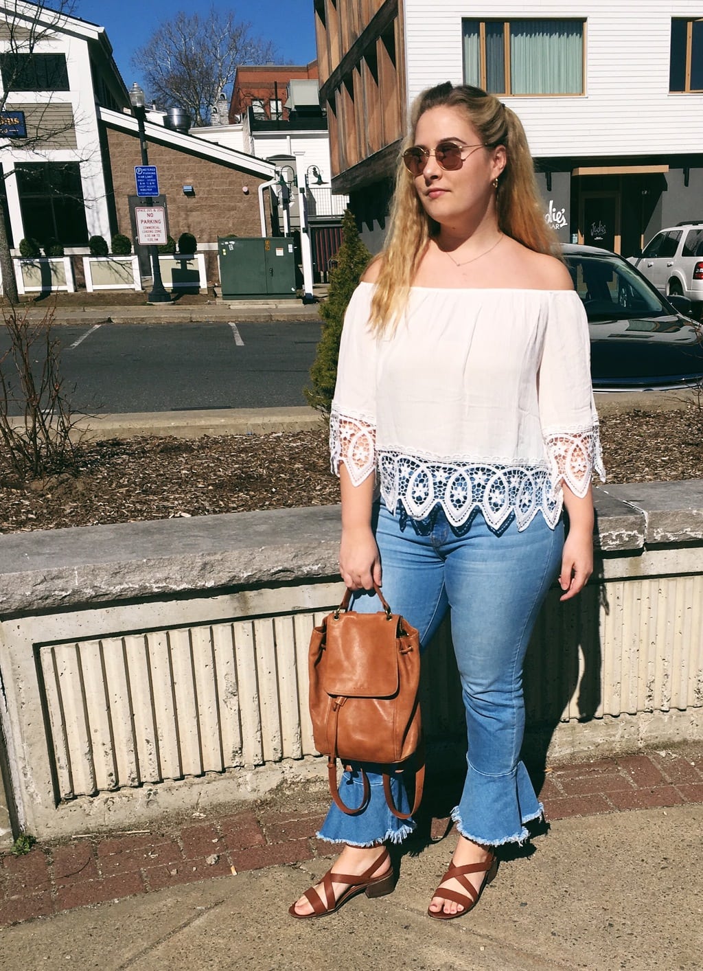 University of Massachusetts Amherst student Kendall wears a cream off-the-shoulder bohemian top with lace detailing on the sleeves and hem with high-waisted light-wash denim cropped flare jeans, a leather bucket backpack, and brown leather strappy sandals with chunky heels.