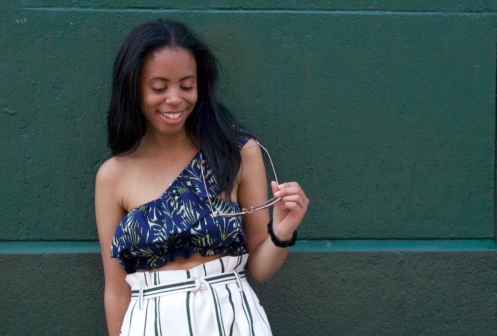 College student fashion: Notre Dame student wears a vertical striped high waisted white mini skirt and a navy and green one-shoulder ruffle crop top and holds a pair of sunglasses