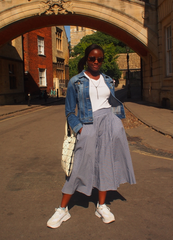 University of Oxford student Chelsea wears a vintage white v-neck tee with a blue and white plaid midi skirt, denim jacket, and white chunky sneakers.