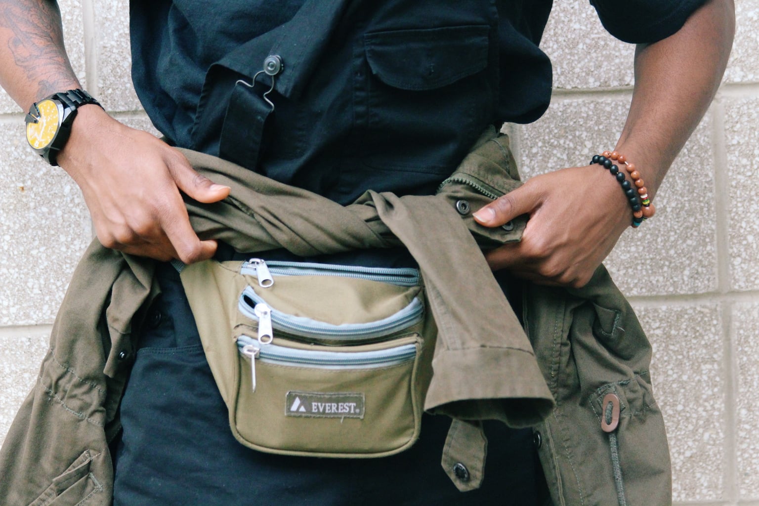 Karon wears a light brown fanny pack with grey zippers and an olive anorak jacket. He accessorizes with black and orange beaded bracelets.