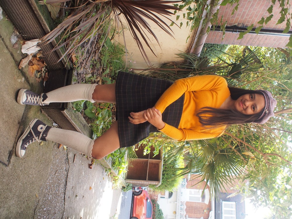 University of Westminster student Sharen wears a mustard yellow-orange long-sleeve v-neck sweater with a high-waisted plaid a-line schoolgirl skirt, a purple hat, and ruffled knee-high socks with low-top Converse sneakers.