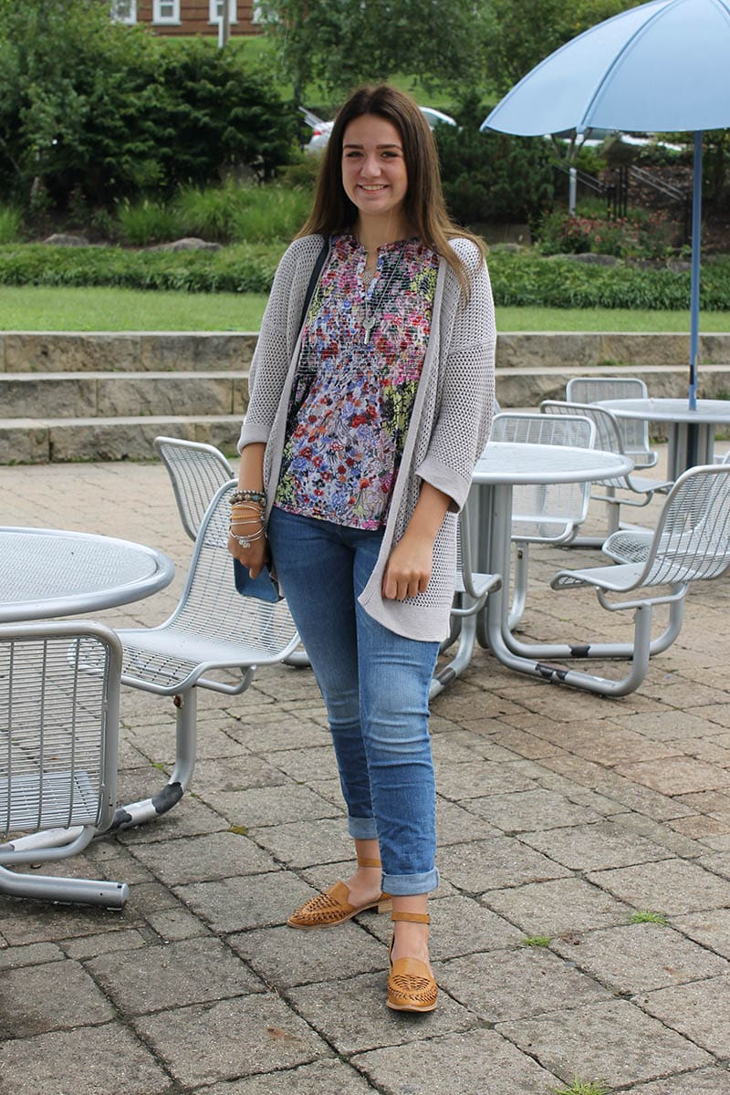 West Virginia University student Alyssa wears a blue, pink, and green loose floral blouse with an open-weave grey knit cardigan, mismatched jewelry, casual denim jeans, and loafer-style sandals.