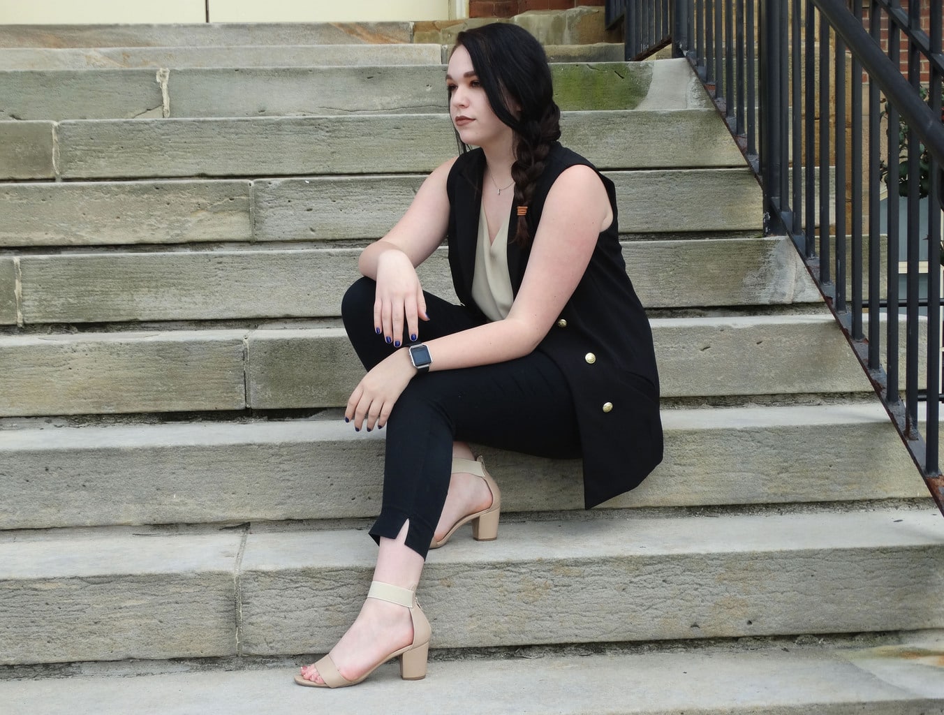 Bree wears a black military-inspired vest with gold buttons, a tan v-neck tank top, and black capri trousers with tan chunky-heeled sandals.
