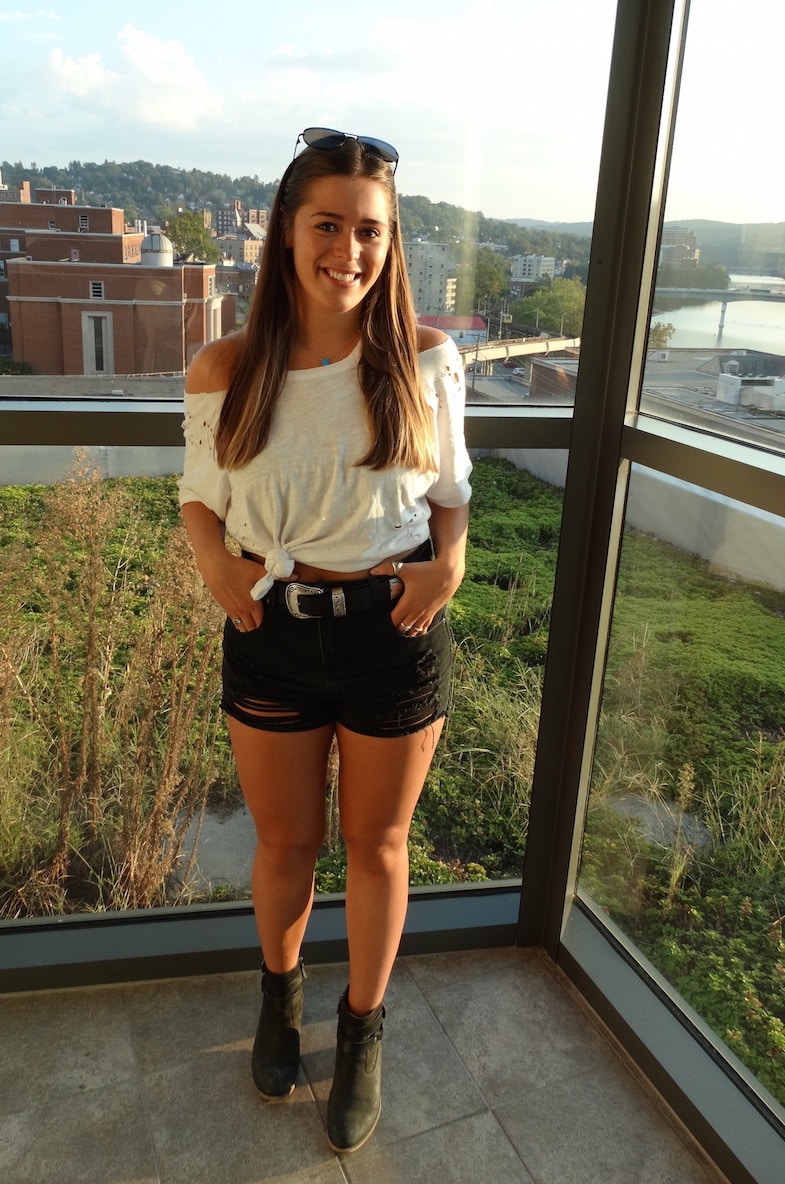 Carly, a student at West Virginia University, wears a distressed white off-the-shoulder tee tied to show off her high-waisted black denim shorts and cowboy-inspired belt buckle.