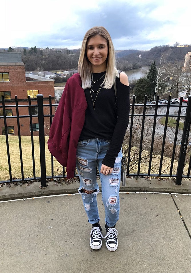 West Virginia University student Courtni wears a black cold shoulder long sleeve sweater with light-wash shredded distressed denim jeans and fishnets underneath them. She finishes off her look with classic black Converse sneakers, a gold layered necklace, and a magenta bomber jacket.