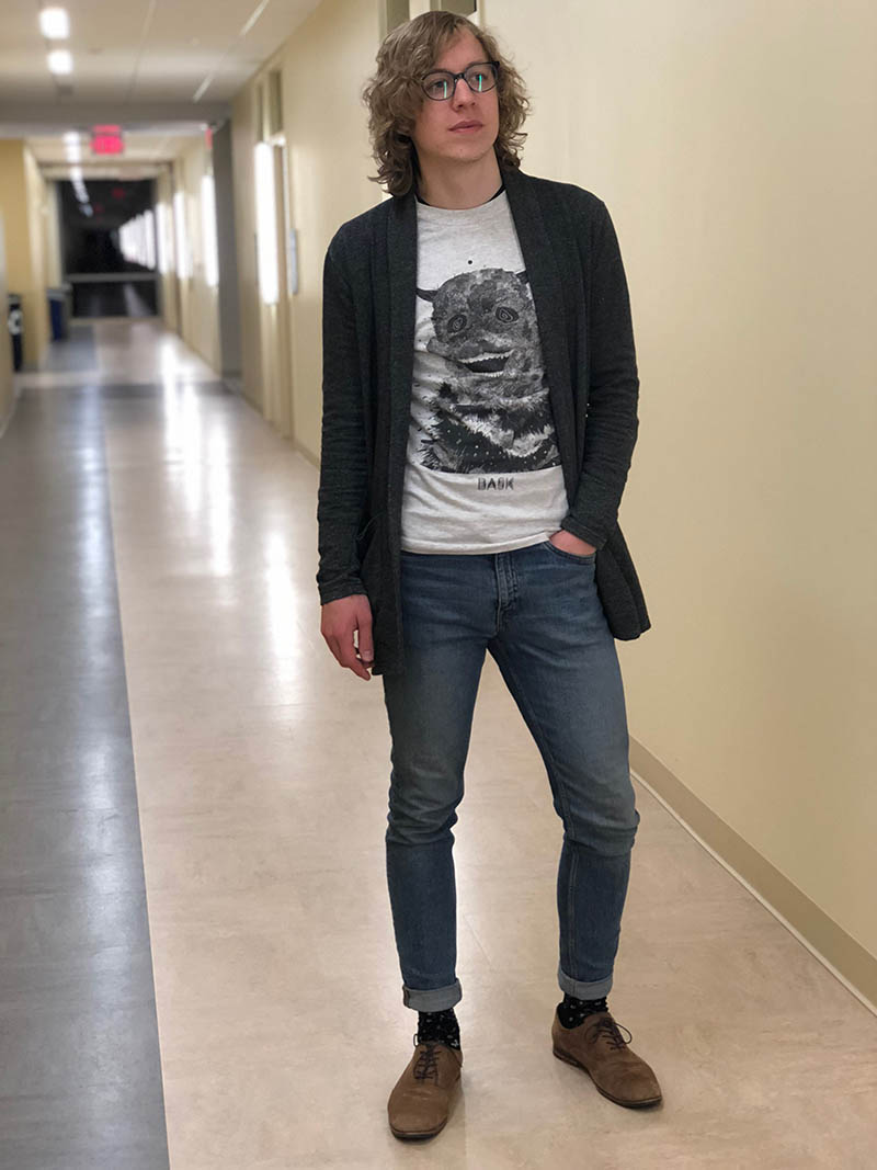 West Virginia University student Davey sports a light grey graphic tee with a dark grey knit cardigan with pockets, fitted and cuffed jeans, black dress socks, and flat brown lace up loafers.