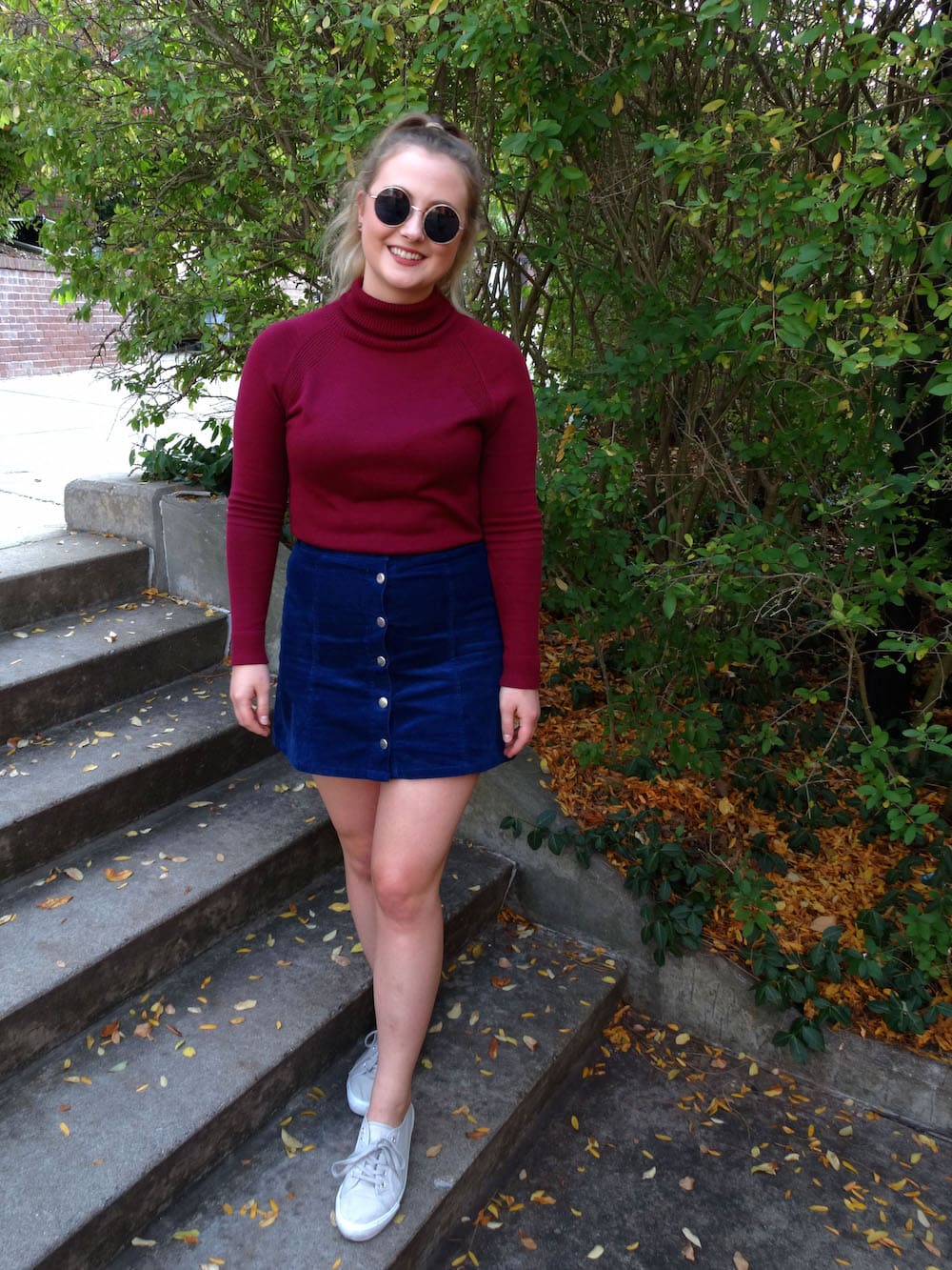 West Virginia Student Emma in a fall-forward outfit. She pairs a maroon turtleneck sweater with a high-waisted blue button-up corduroy miniskirt with grey sneakers and circular frame sunglasses.