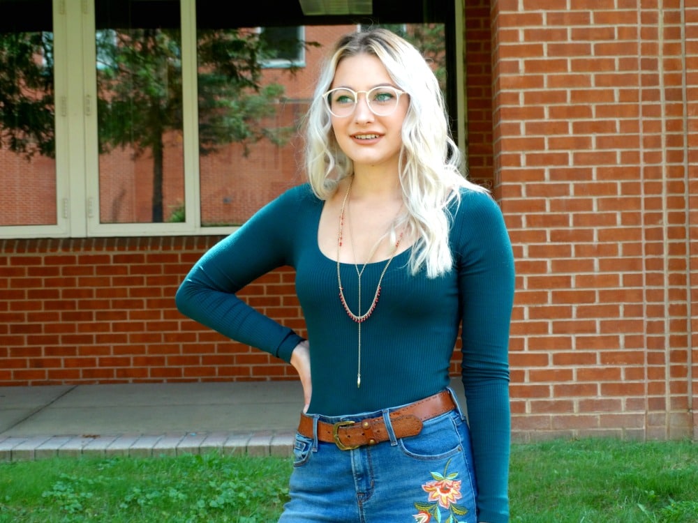 West Virginia University student wears a scoop-neck long-sleeve teal top tucked into a pair of embroidered high-waisted jeans with a chunky brown belt and layered gold necklaces.