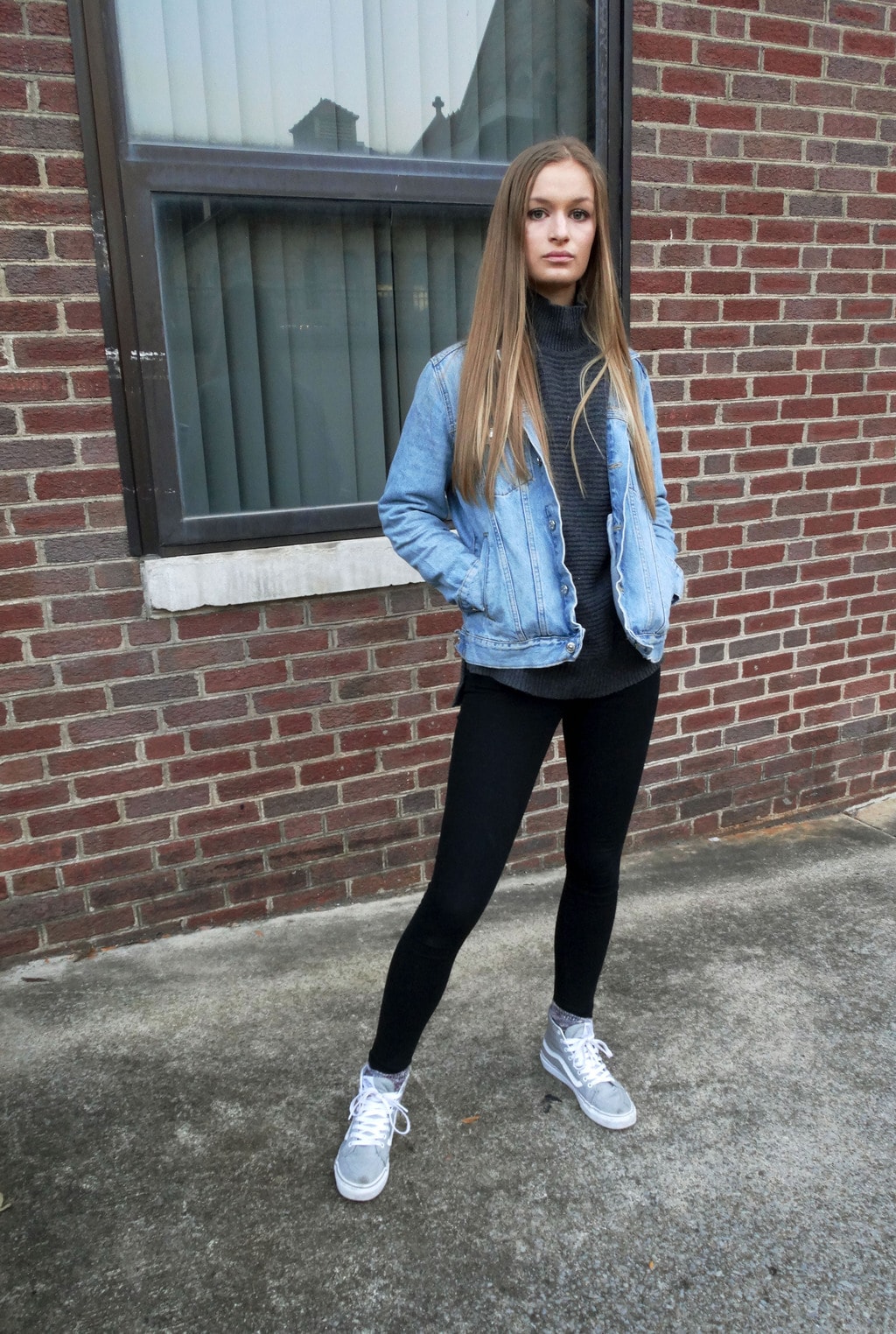 This student wears a space grey turtleneck sweater with an oversized, distressed denim jacket, black leggings, and sporty grey sneakers with white detailing.