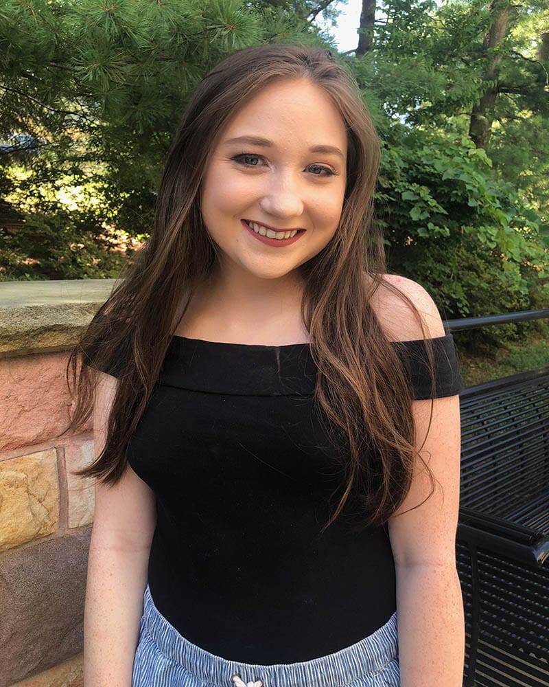 Maggie wears a black off-the-shoulder fitted top tucked into her pants.