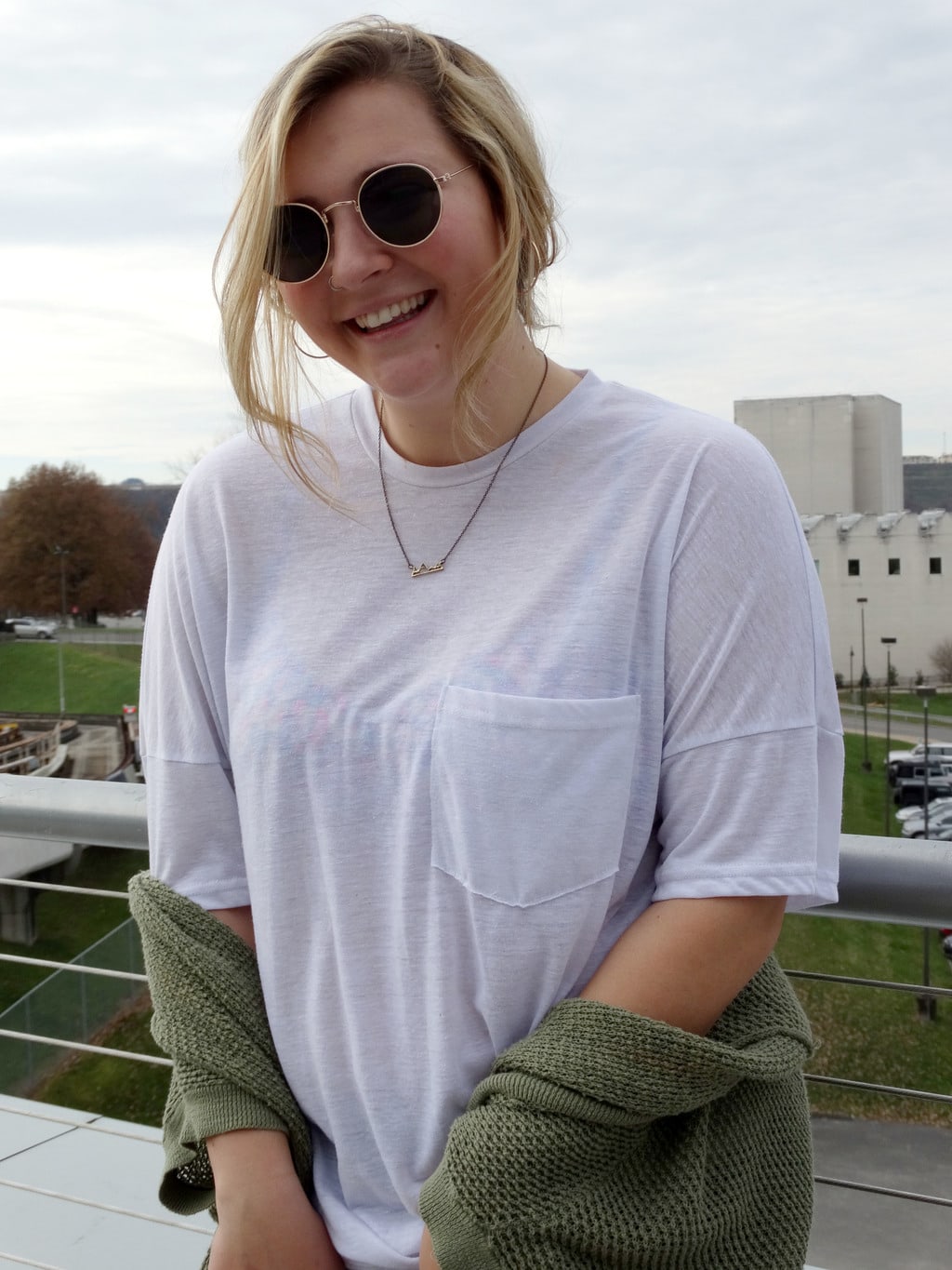 Mel wears a loose-fitted white pocket t-shirt with a simple gold necklace and round sunglasses with gold frames.