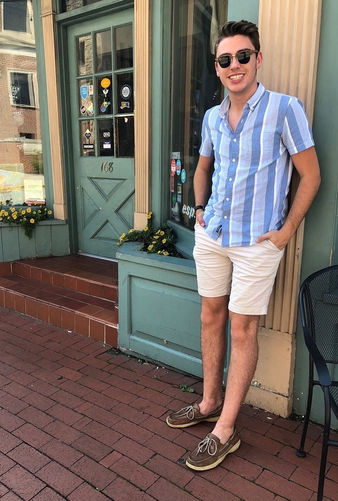 Nathan wears a summery collared button-up with various shades of blue and white vertical stripes, Ray-Ban sunglasses, khaki shorts, and brown leather Sperry boat shoes.