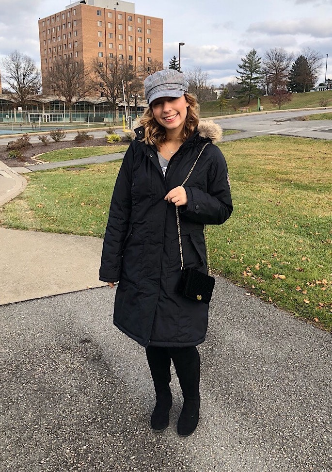 West Virginia University student Sammie wears a long navy coat with a faux fur lined hood, tall black boots, a grey plaid cabby hat, and a black purse with a gold chain strap.