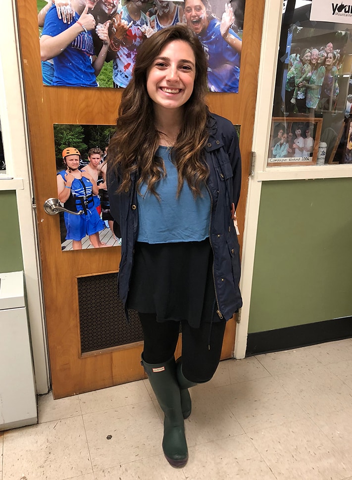 West Virginia University student Sarah is rainy day ready in an azure blue crop top with lace detailed sleeves, a high-waisted black flowy miniskirt, opaque black tights, and iconic hunter green rain boots. She also wears a blue trench coat with gold accents.