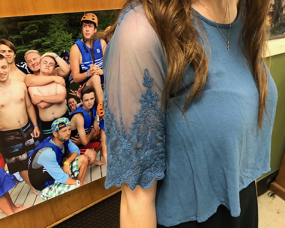 Sarah sports a light blue crop top with sheer sleeves that have lace detailing and a simple gold cross necklace.