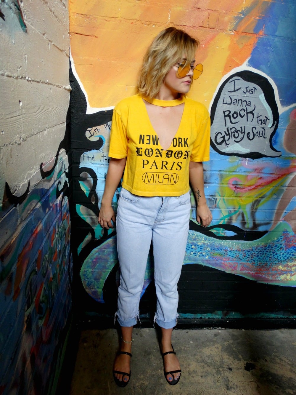 Fashion at West Virginia University: Student Shelby rocks a trendy outfit consisting of a yellow v-neck tee with cutout detail that reads New York Paris London Milan, light wash mom jeans, black sandals, and round yellow sunglasses