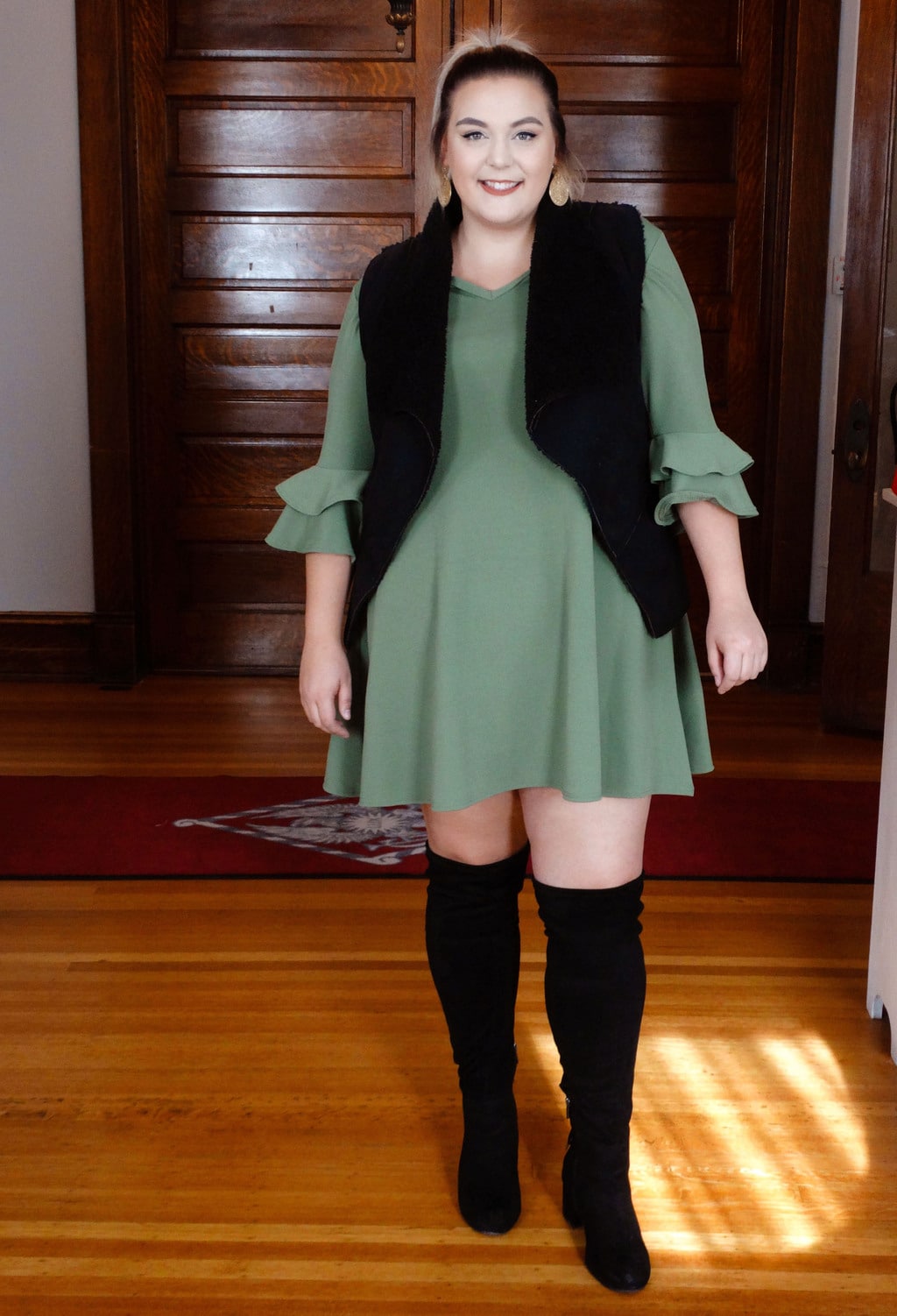 Sydney wears a green dress with layered ruffle bell sleeves and a black furry vest with black over-the-knee boots.