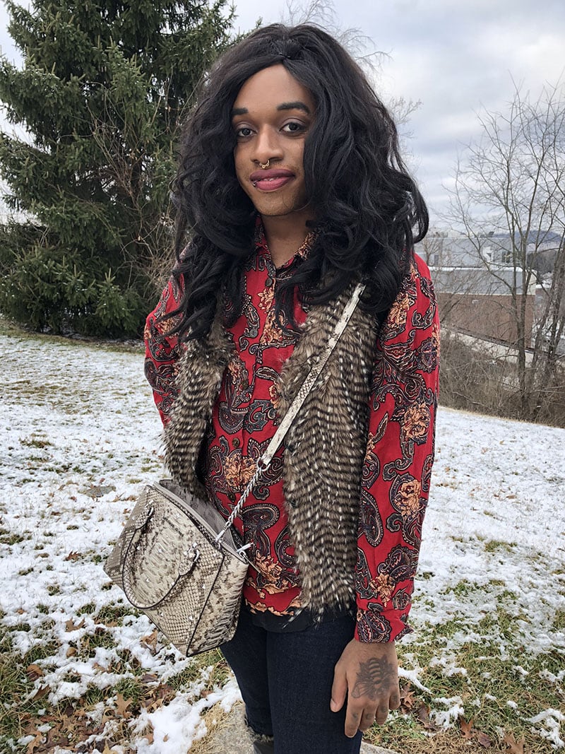 Teddy wears a bright red button-up blouse with a loud paisley print. He pairs it with a brown and black striped faux fur vest and a Michael Kors cream snakeskin crossbody purse.