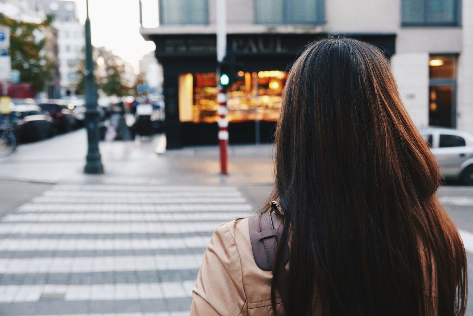 Photo of brunette girl wearing a tan jacket walking down a city street.