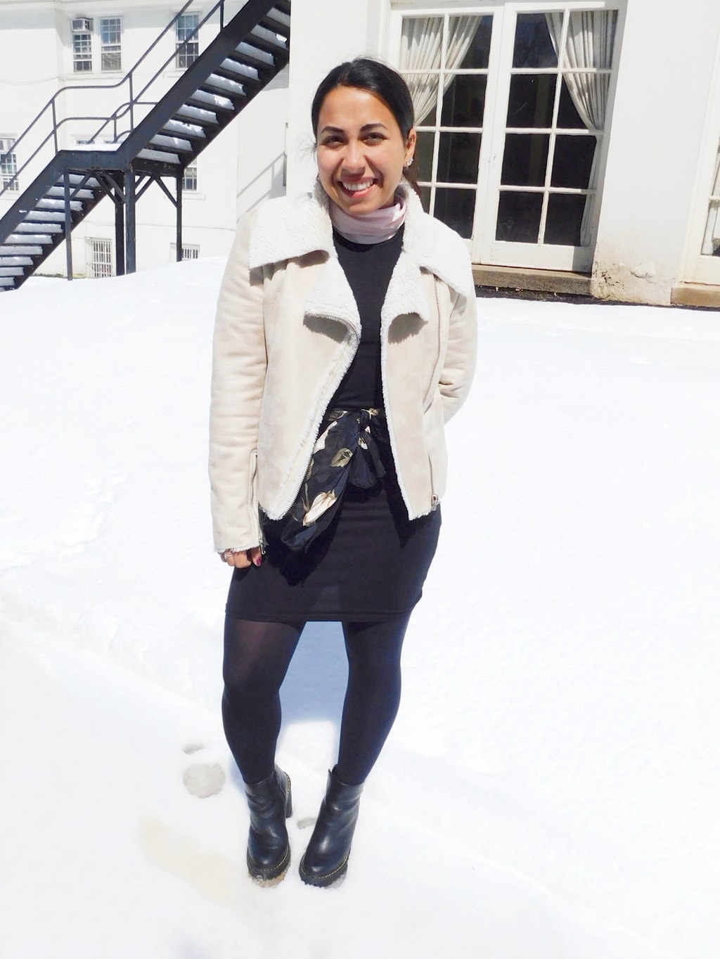 Jefferson University student Vanessa wears a springtime tank dress with a pastel pink long sleeve turtleneck underneath, opaque black tights, a white motorcycle style shearling-lined coat, a feminine black and pink scarf worn as a belt, and black rubber Doc Martens booties.