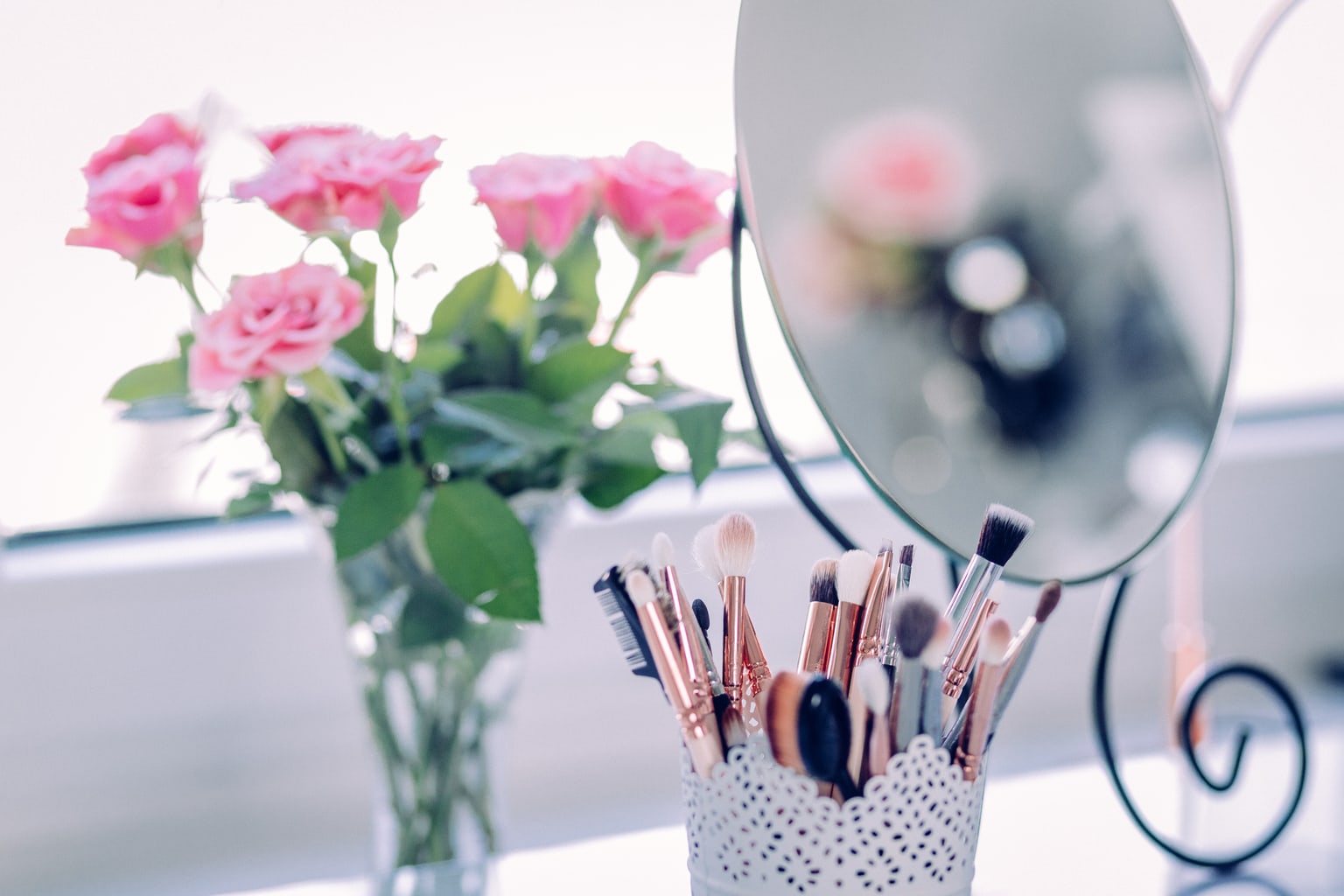 Makeup brushes in a lace pencil holder on a vanity, behind a mirror and pink flowers