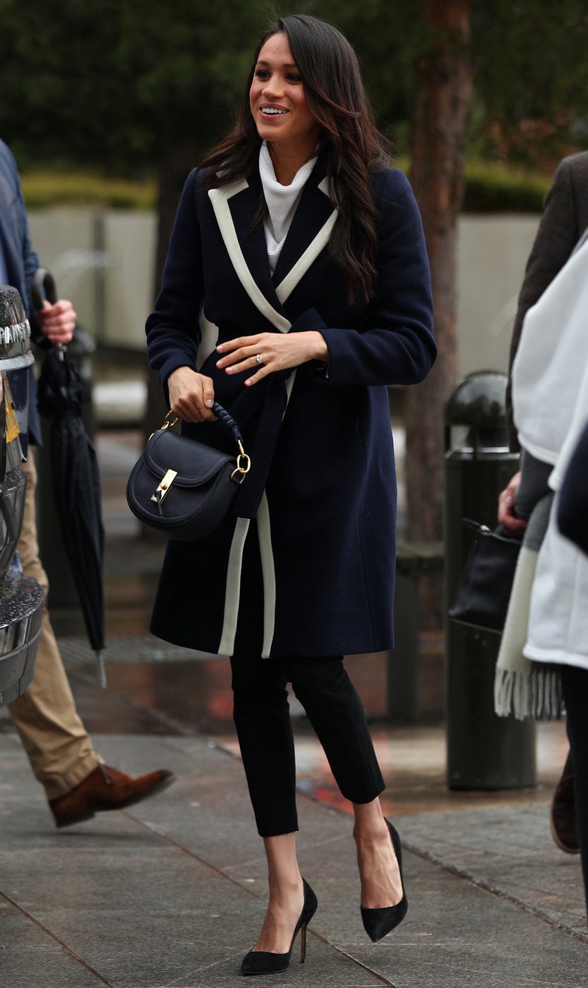 Meghan Markle wearing a navy blue coat with white collar lapel detailing, a white turtleneck sweater, simple black pumps, and a black and gold handbag