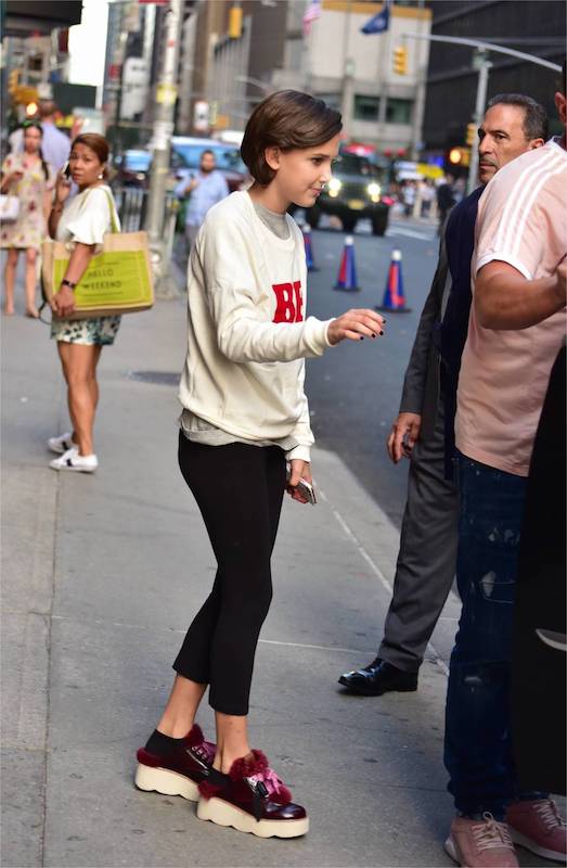 Millie Bobby Brown wearing a graphic print Be Nice sweatshirt, black cropped pants, and platform sneakers with fur accent