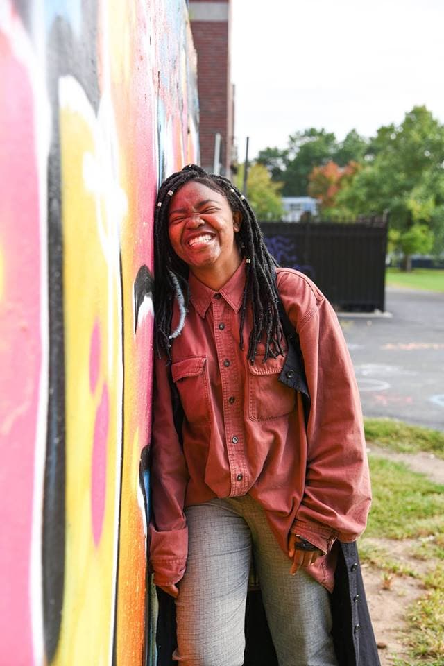 College student wearing an oversized rust colored button down shirt, braided hair, and plaid vintage pants