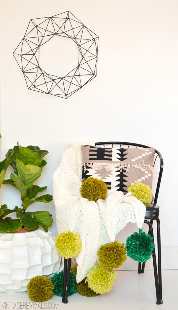 Photo including a chair with a pom pom blanket in it as well as a pillow. There is a plant to the left of the chair and a metal wall hanging above it.