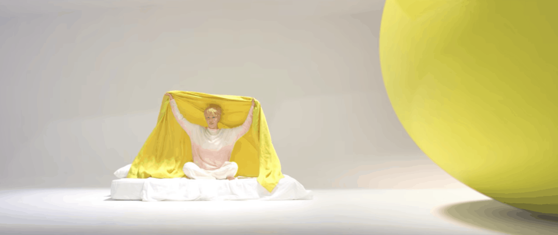 BTS Serendipity music video: Band member on a white floor covered with a bright yellow sheet, sitting next to an oversized yellow ball