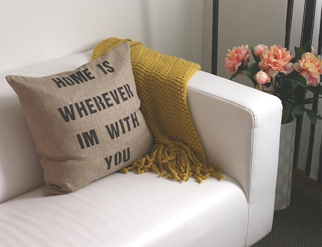 Photo of a stenciled pillow on a white couch with a yellow blanket on it. There is a vase of flowers next to the couch.